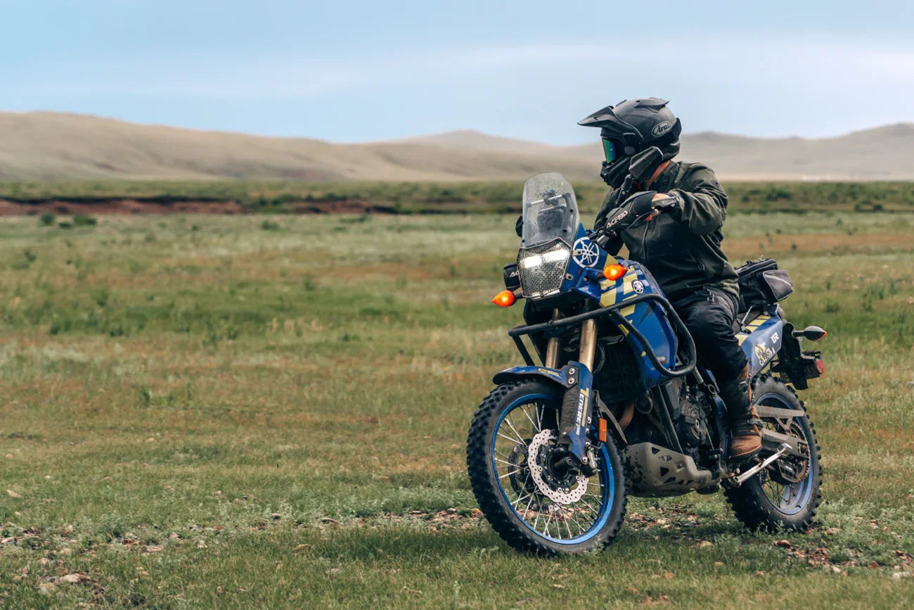 Rider in black Arai helmet on blue adventure motorcycle in open grassland, showcasing protective gear and off-road readiness.